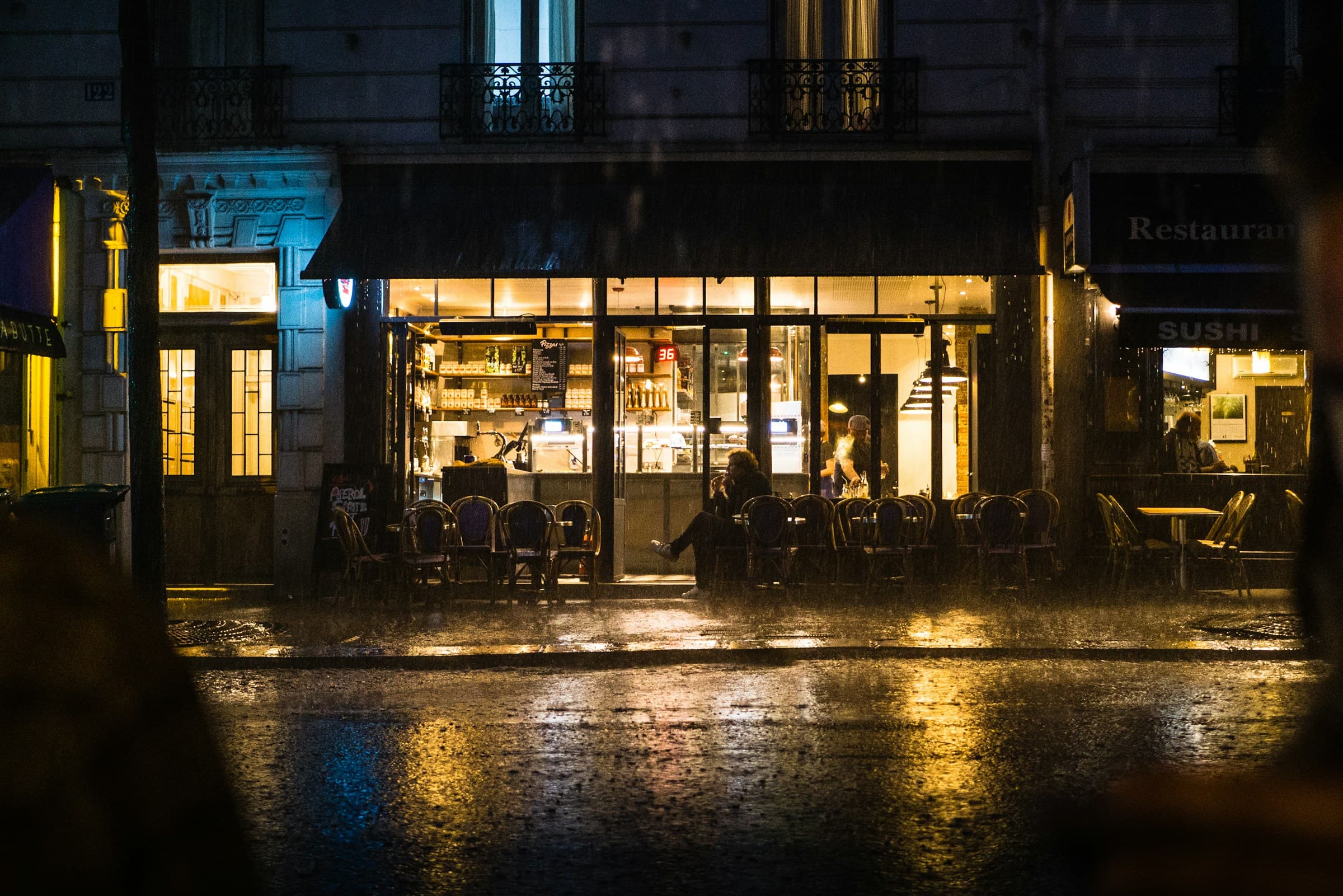 Interiorul unui bar mic, seara, lumină caldă, ploaie la geam